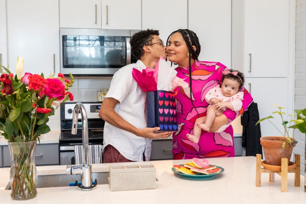 image of a man kissing a woman holding a baby girl, while he's holding a gift bag with hearts on it.