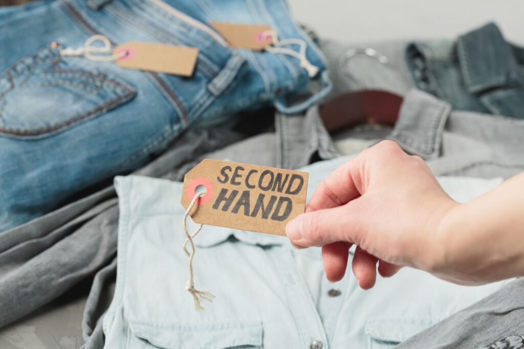 image of a hand holding up a tag that says "second hand" in front of a jean shirt and jeans